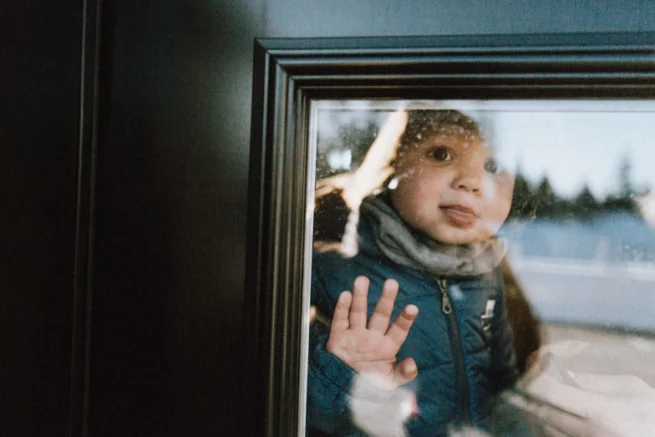 "Photographie documentaire d'un enfant regardant par la fenêtre, capturée en hiver par le photographe 5Mphotographie à Rimouski. L'image met en lumière la spontanéité et l'émotion d'un moment intimiste, avec la neige en arrière-plan et une lumière naturelle douce, typique d'une séance photo hivernale."
