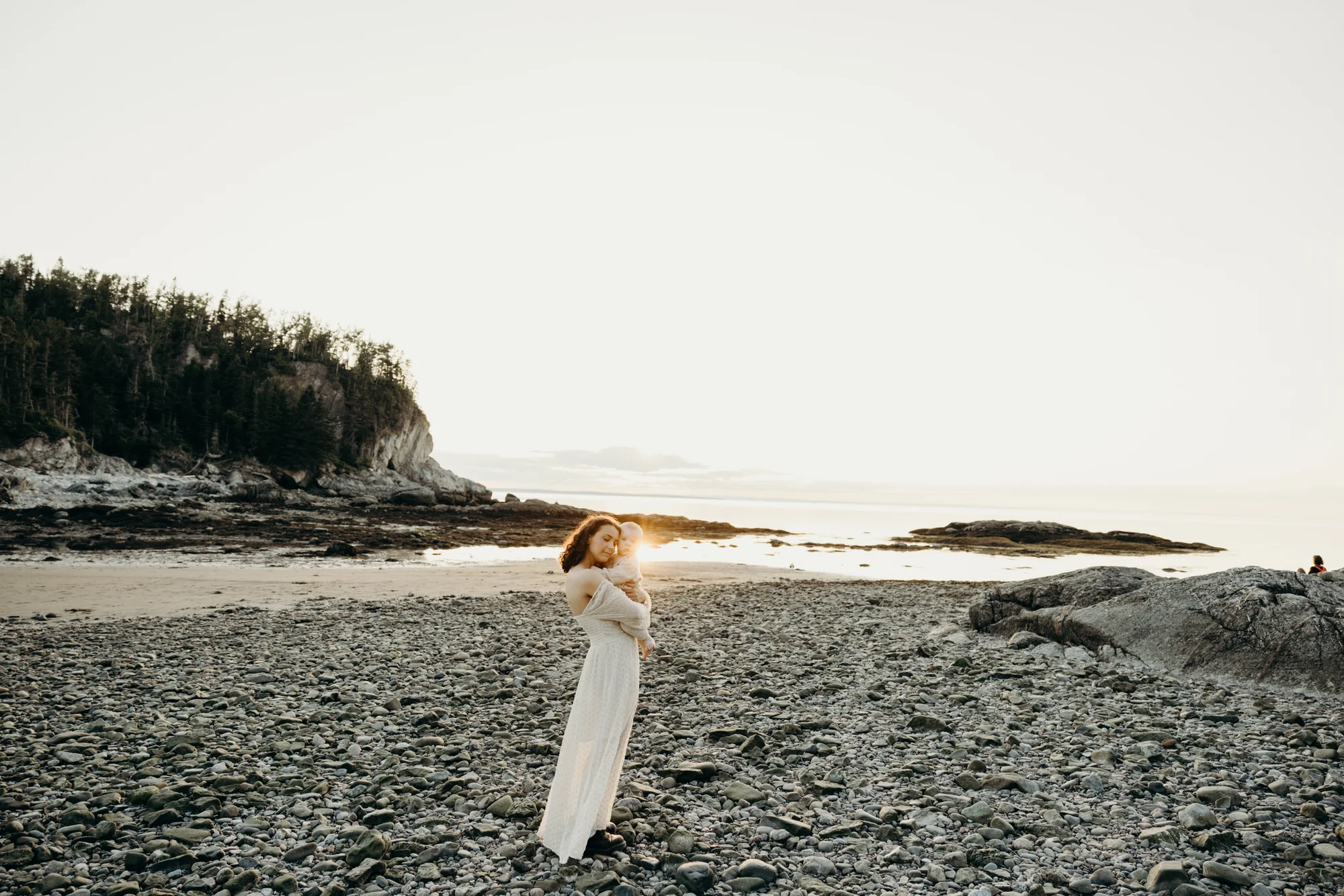 Future maman en robe longue blanche debout sur la grève de galets, séance maternité en lumière naturelle.