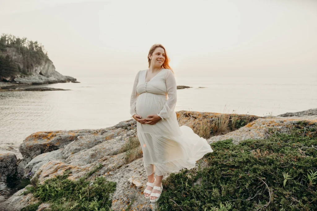 Femme enceinte tenant son ventre et souriante avec une robe blanche qui vole au vent prise par 5Mphotographie.