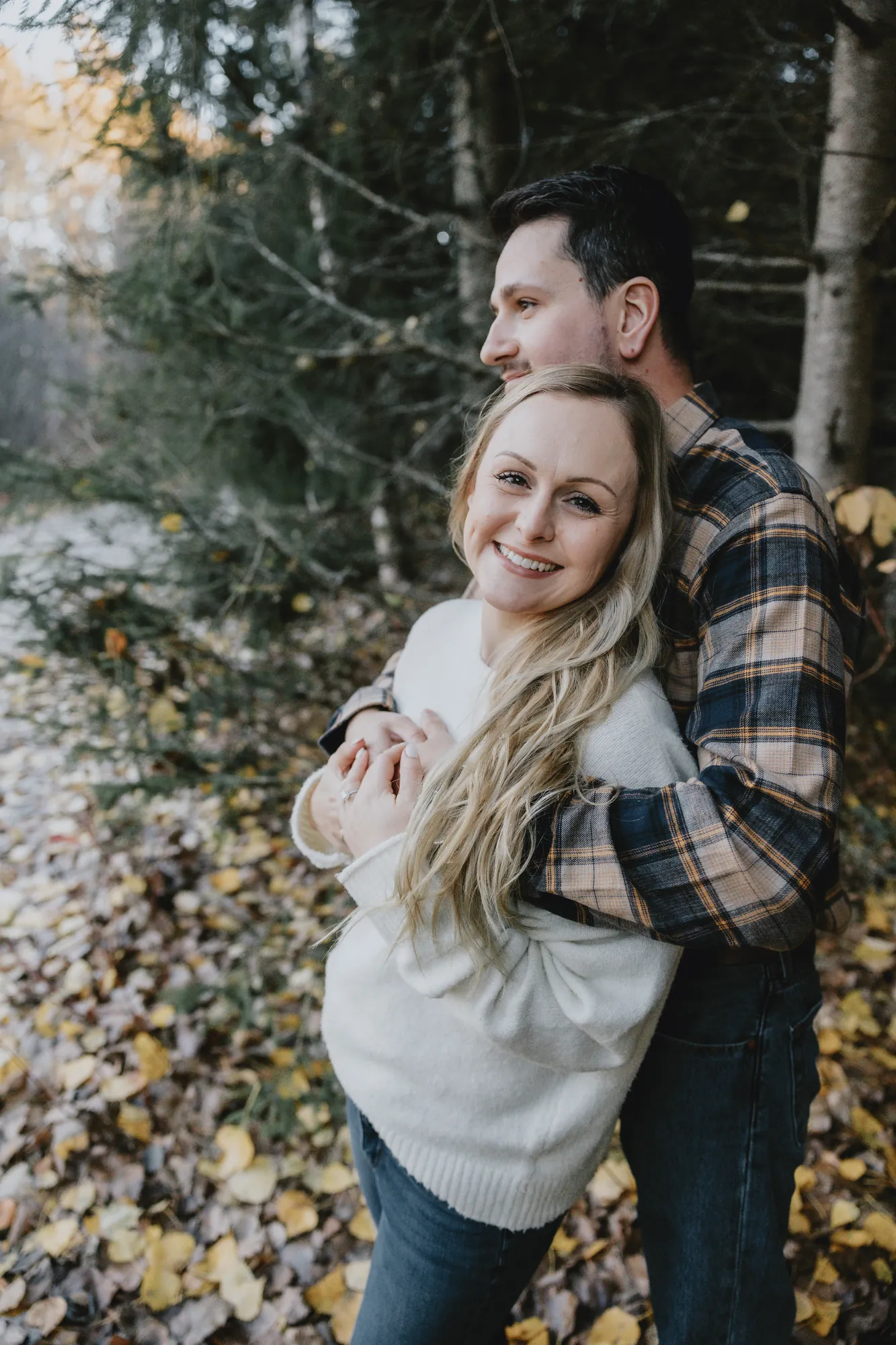 Nathalie souriante dans les bras de Guillaume près du bois.