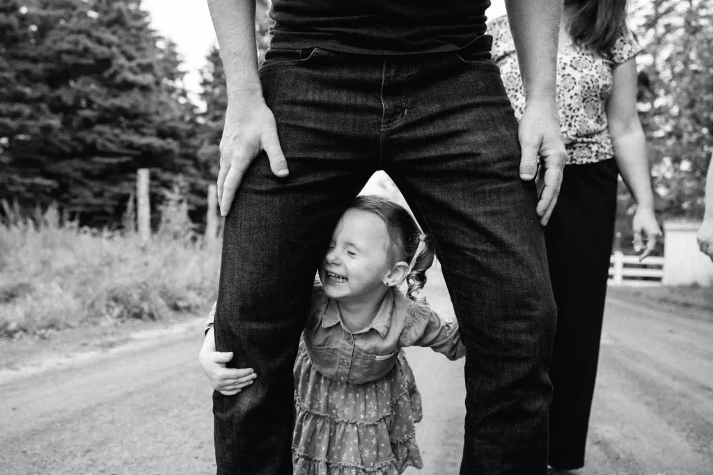 Photo noir et blanc d'une petite fille qui se cache sous les jambes de son père en riant. Photo prise par 5Mphotographie.