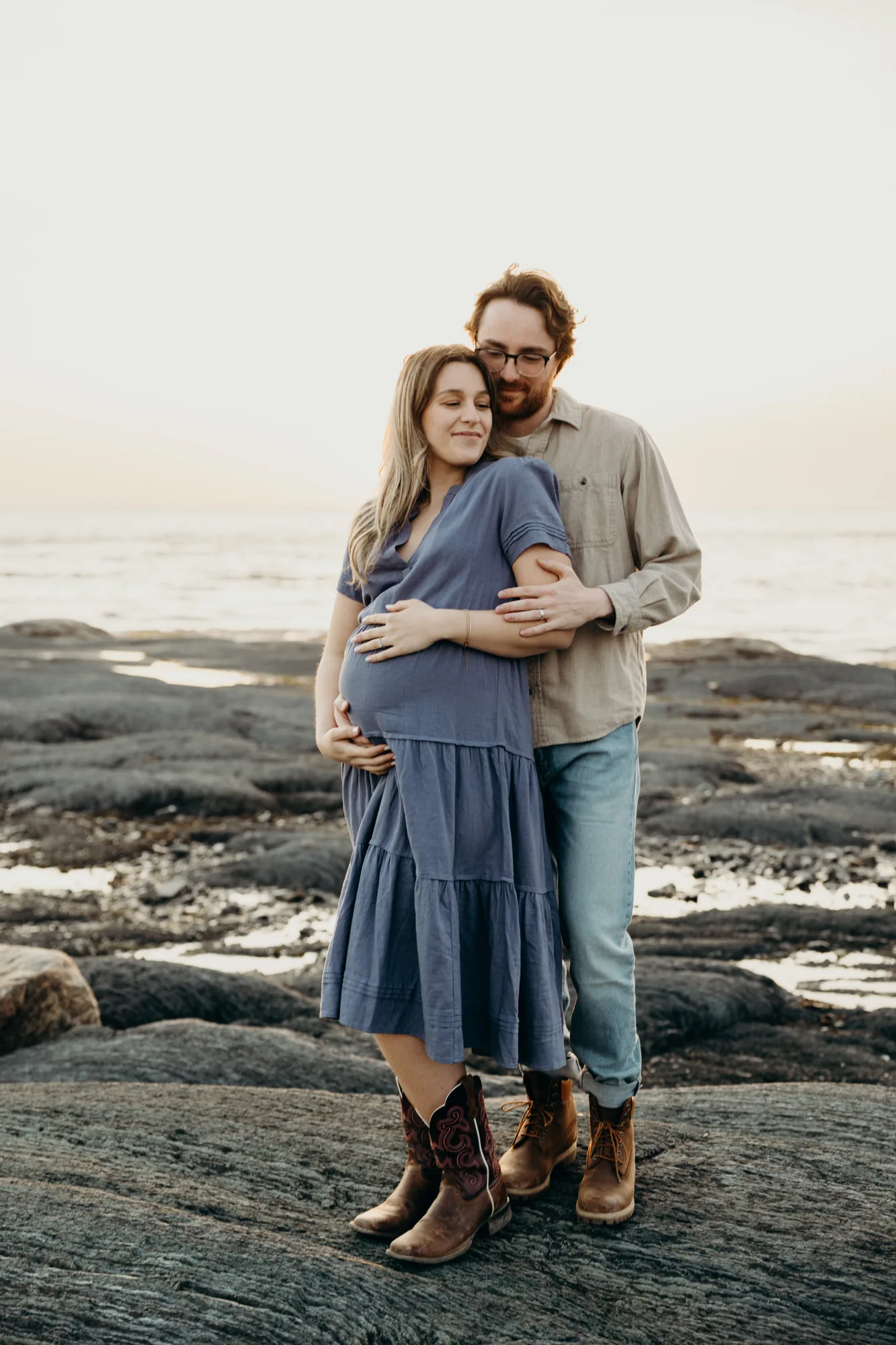 Future maman et son conjoint en robe bleue posant sur les rochers du littoral regardant le coucher de soleil avec bottes de cowboys.