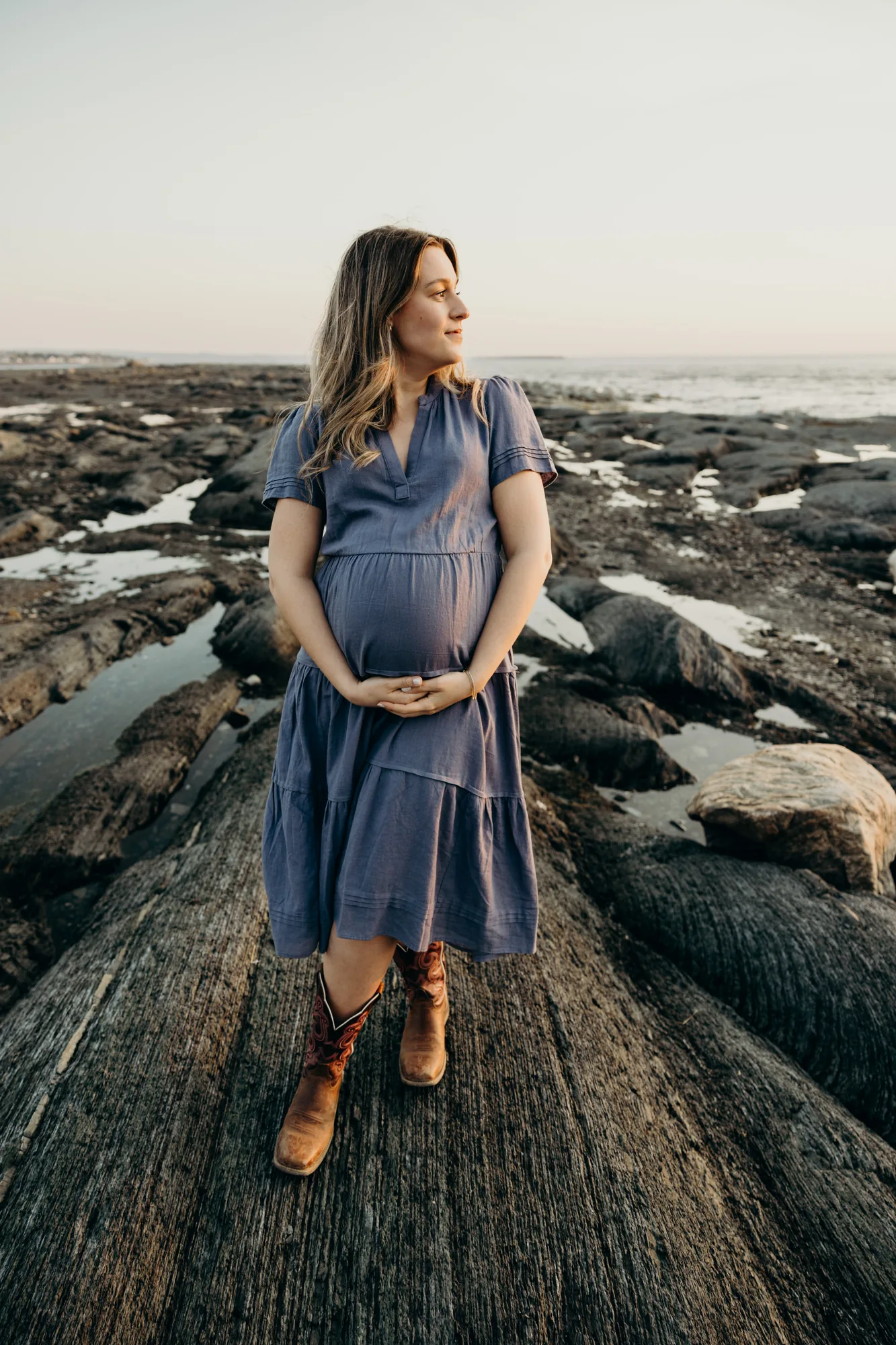 Future maman en robe bleue posant sur les rochers du littoral regardant le coucher de soleil avec bottes de cowboys.