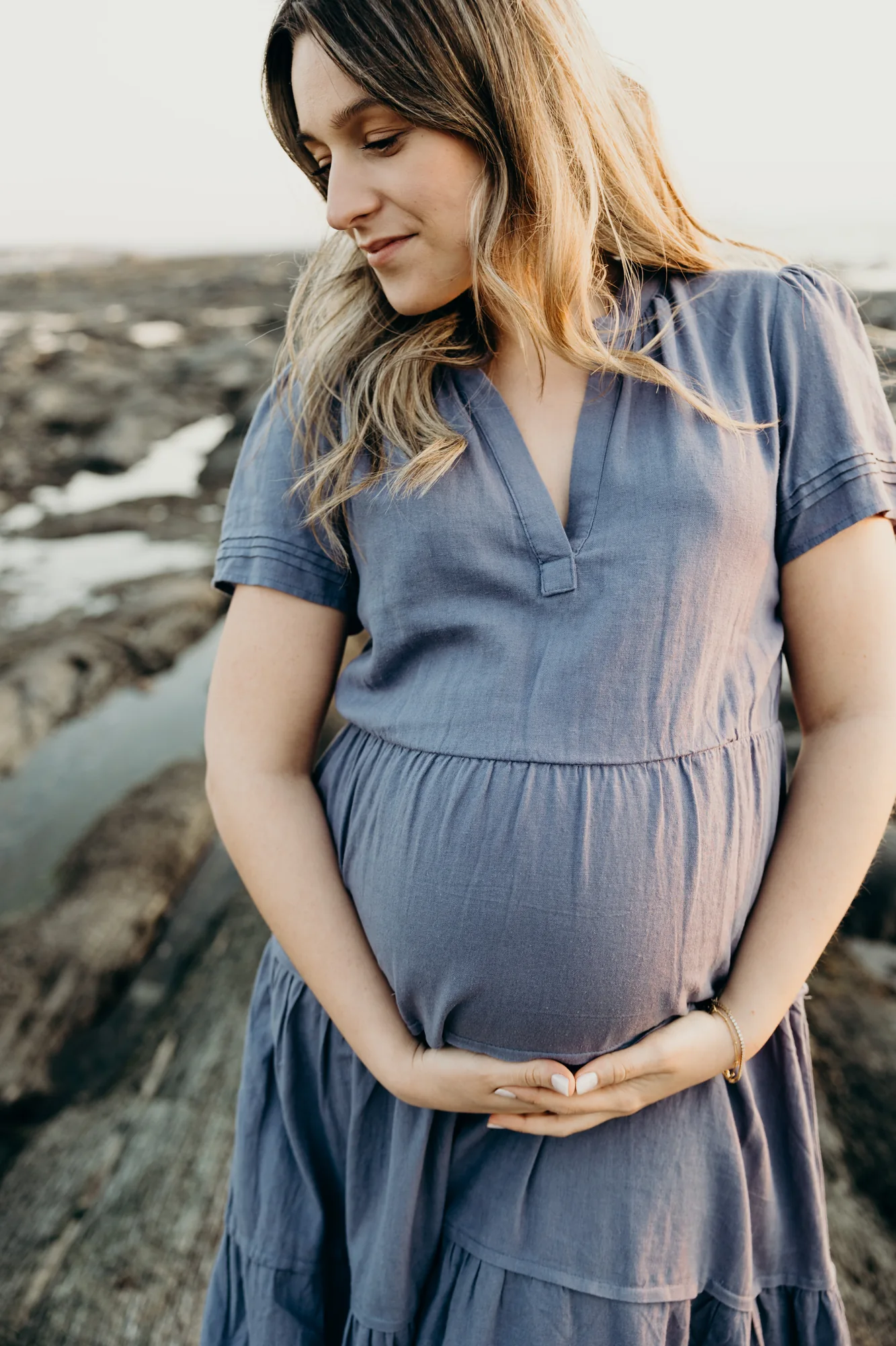 Future maman en robe bleue posant sur les rochers du littoral les mains sur son ventre.