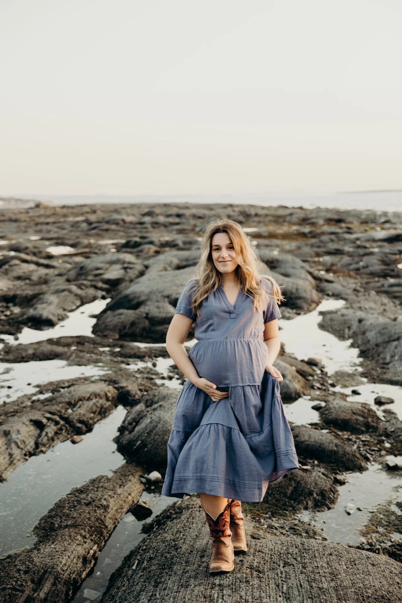 Future maman en robe bleue posant sur les rochers du littoral.